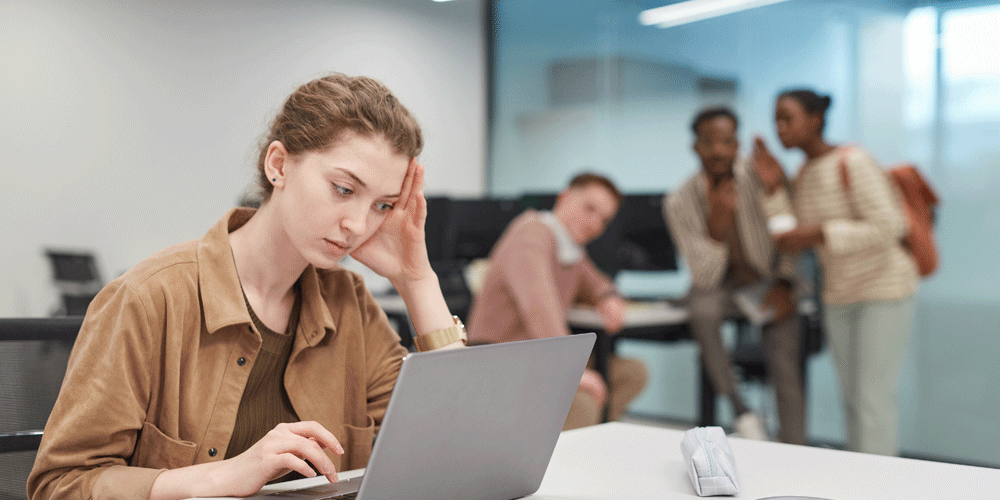 Stressed young woman working on her laptop. A group of coworkers looks on.